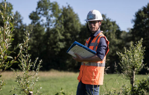 Photo d'un homme avec veste fluorescente, casque de sécurité, avec un porte documents, dans un espace très végétalisé.